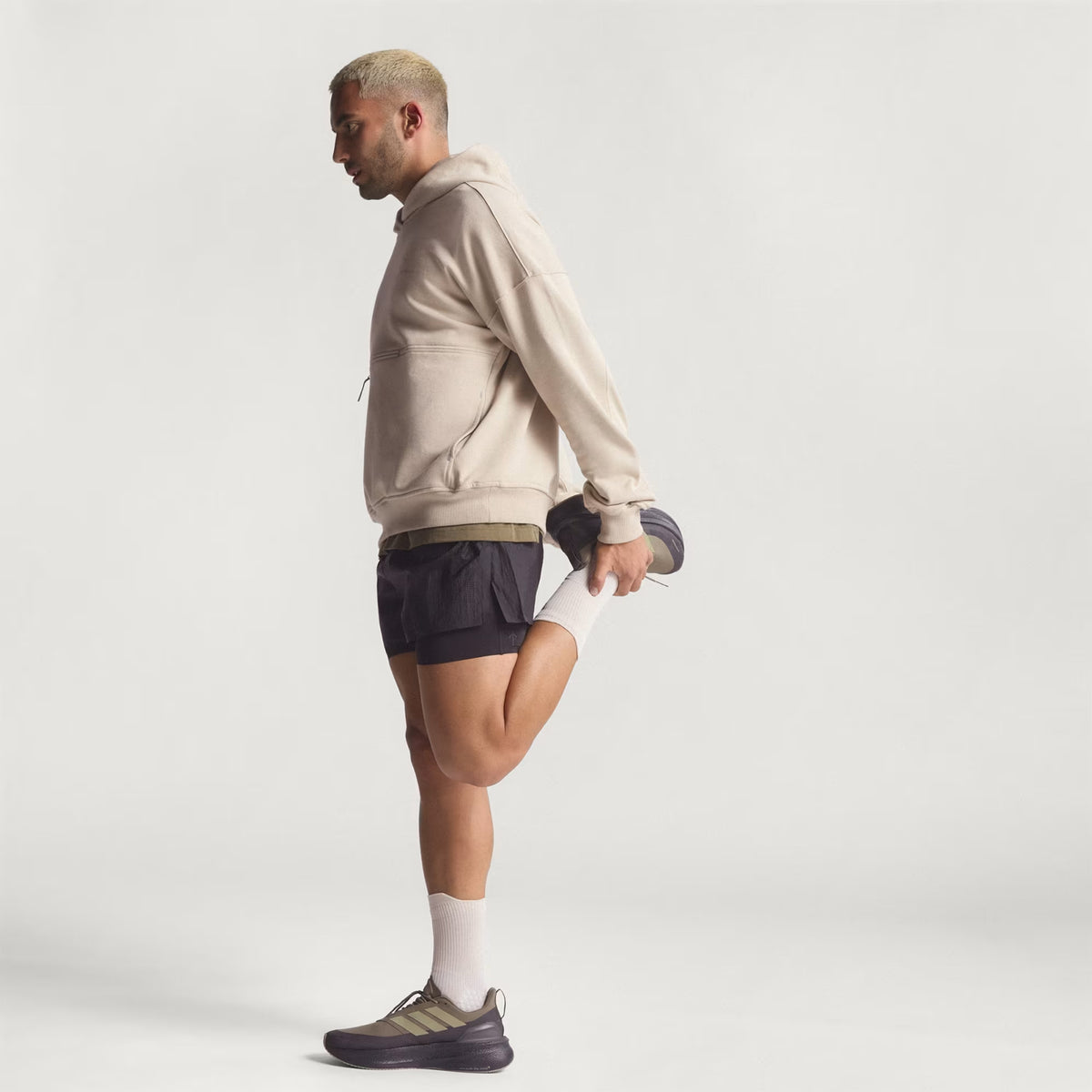A man wearing the Unisex Adi365 H.Koumori Running Hoodie in Putty Grey by Adidas and black shorts stands on one leg, holding his other foot behind him in a quadriceps stretch against a plain white background.