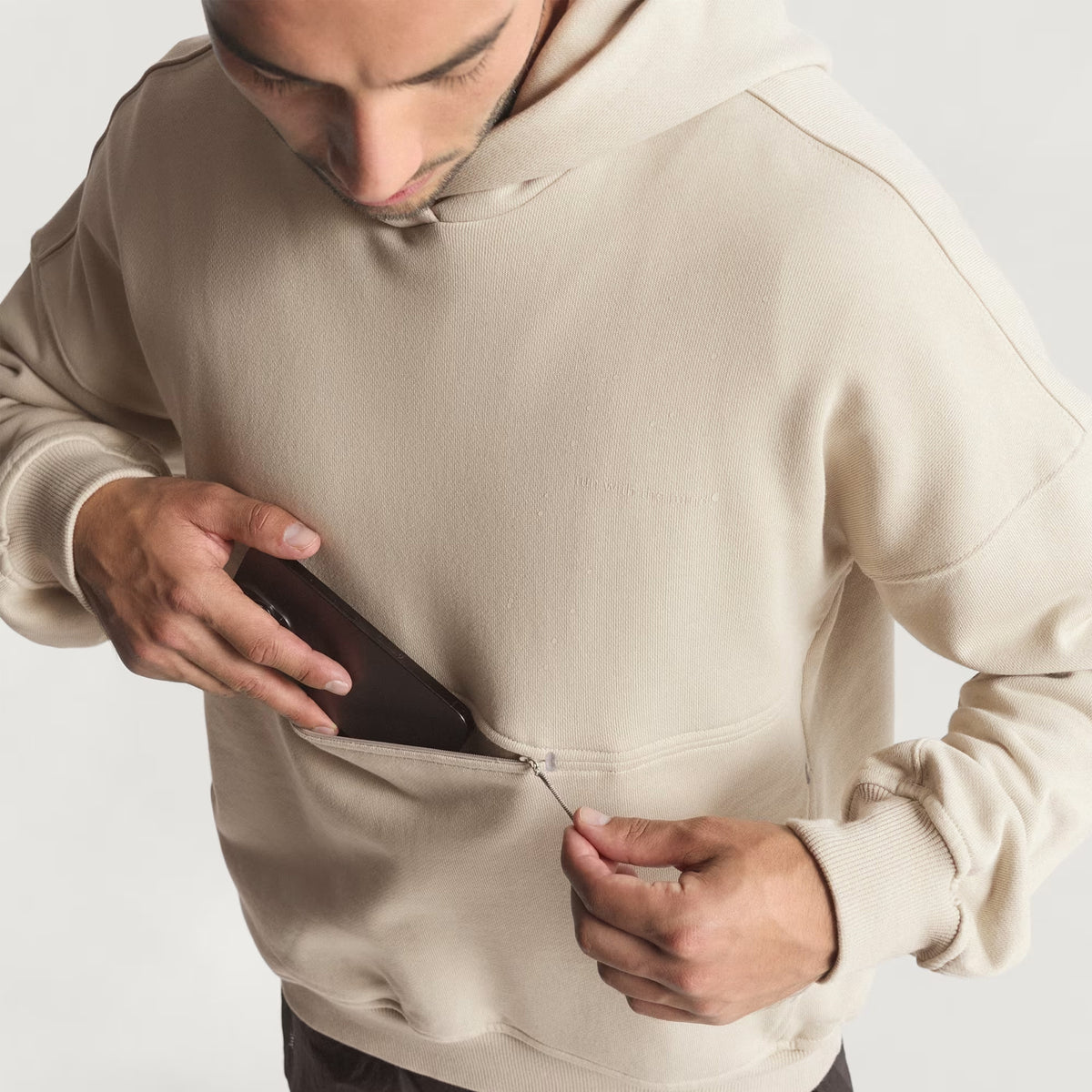 A person wearing the Adidas Unisex Adi365 H.Koumori Running Hoodie in Putty Grey unzips a front pocket to place a black smartphone inside. The image is taken from above, showing only the upper body.