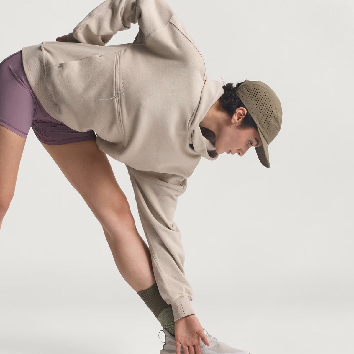 A person wearing the Unisex Adi365 H.Koumori Running Hoodie in Putty Grey by Adidas, along with purple shorts and a tan cap, bends forward to touch one foot while stretching against a plain light background.