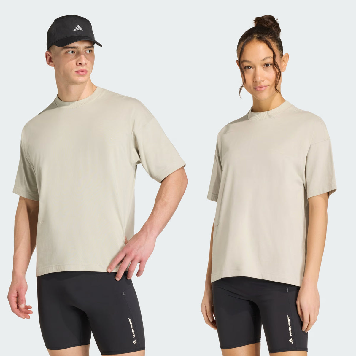 A man and woman wear matching Adidas Unisex Adi365 H.Koumori Running Tees in Putty Grey, paired with black athletic shorts. The man also wears a black Adidas cap, coordinating with his running tee. Both stand facing slightly toward the camera against a plain light background, showcasing their stylish all-gender running shirts from Adidas.