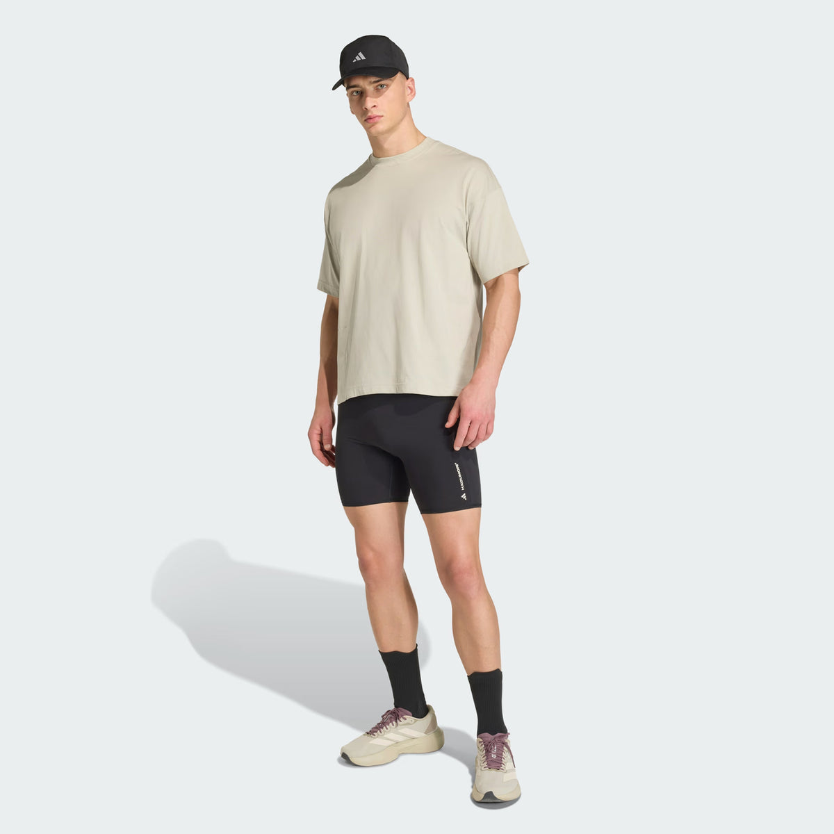 Wearing the Unisex Adi365 H.Koumori Running Tee in Putty Grey from Adidas styled as an oversized T-shirt, the individual pairs it with a black cap, black fitted shorts, black socks, and beige sneakers while standing against a light gray background.