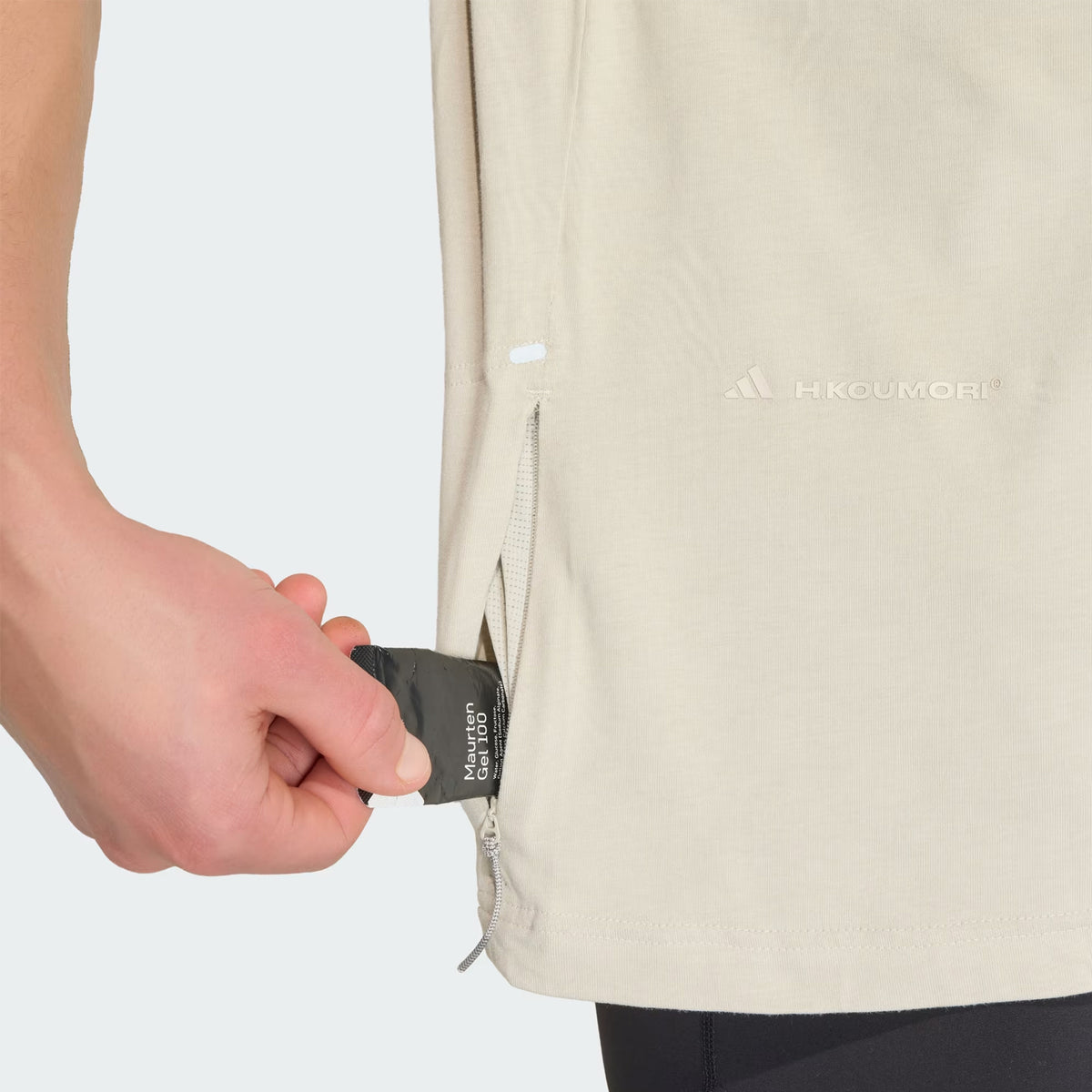 A hand pulls a black tag from the side zippered pocket of the Unisex Adi365 H.Koumori Running Tee in Putty Grey by Adidas. This all-gender running shirt features a subtle logo and is styled here with dark pants.