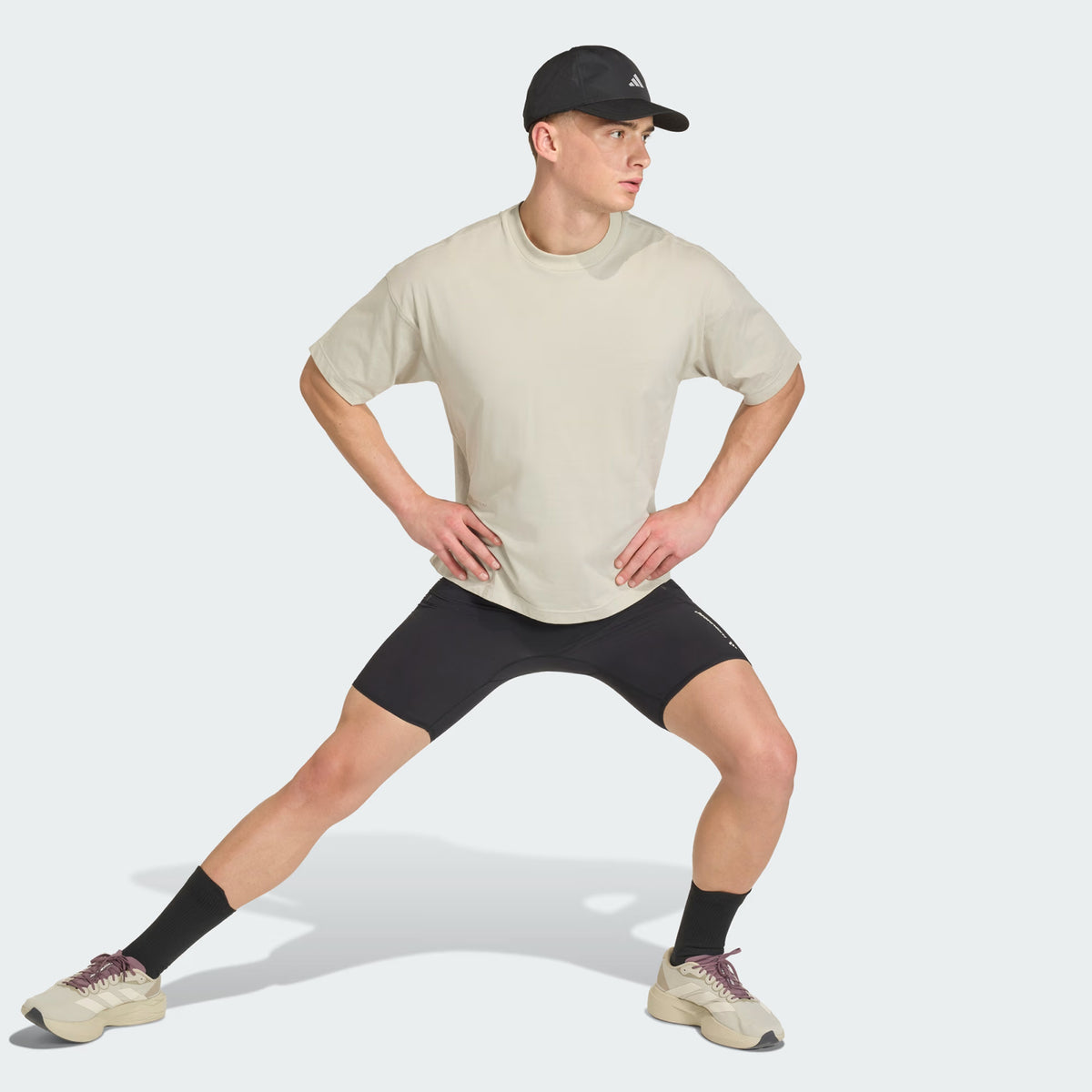 A man in a beige t-shirt, black shorts, black cap, and sneakers performs a side lunge exercise with hands on hips against a plain background.
