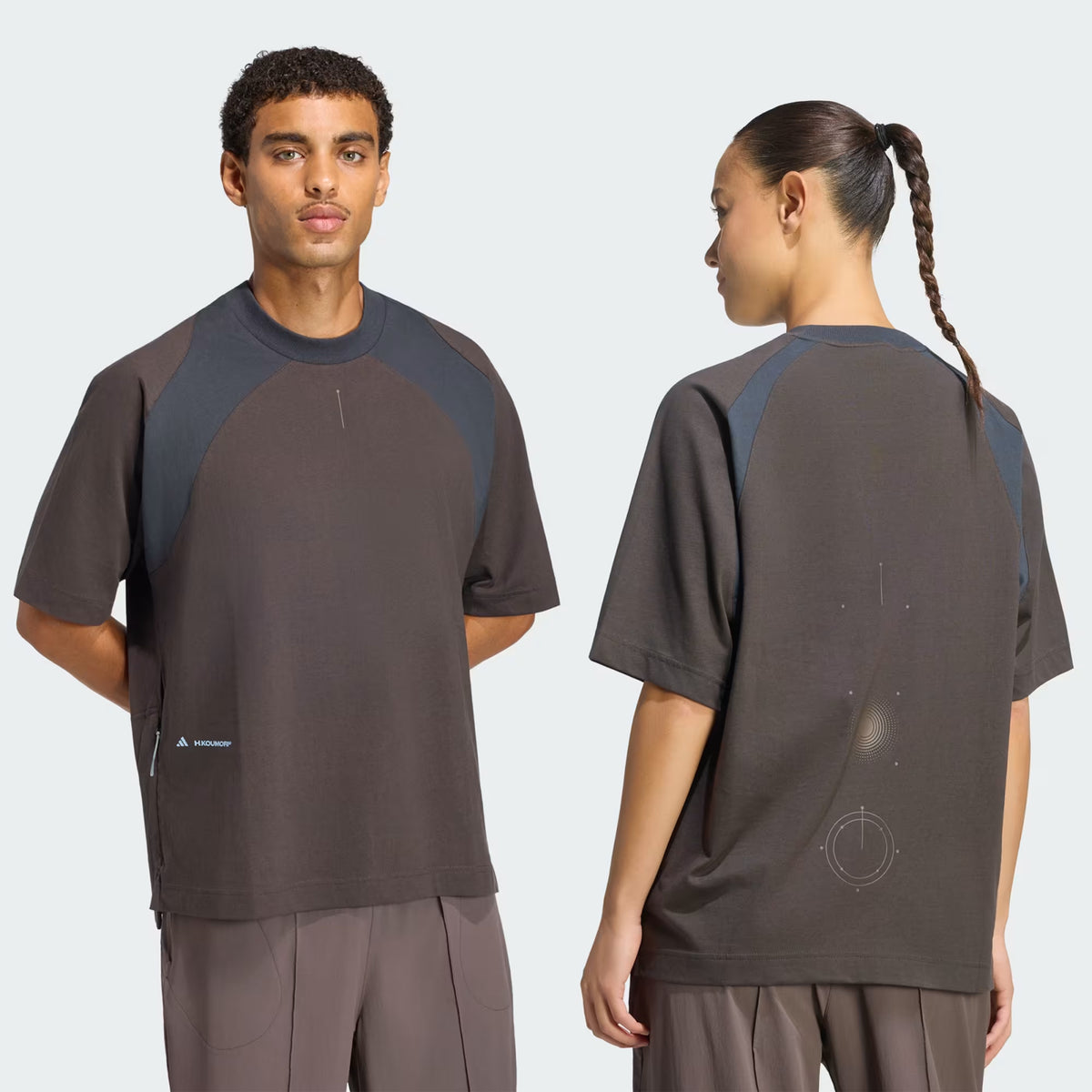A person wears a Unisex H.Koumori Tee Night Brown / Night Grey adidas running tee in brown and dark grey, featuring shoulder paneling and a geometric graphic on the back.