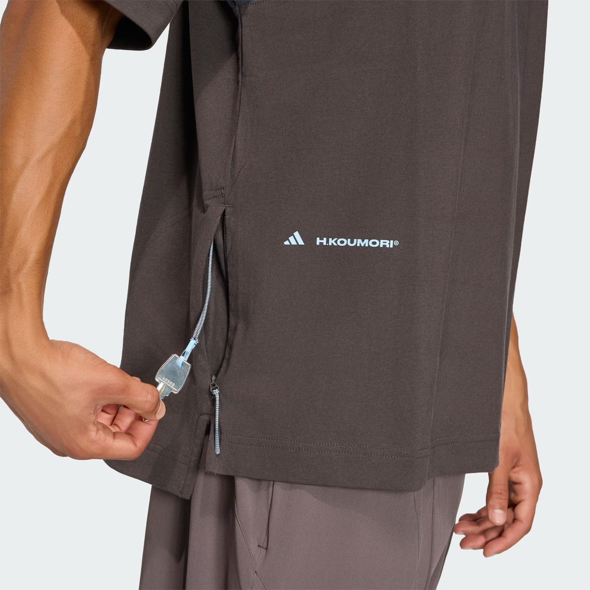 A person pulls the zipper on the side seam of a brown Unisex H.Koumori Tee Night Brown / Night Grey adidas running tee.