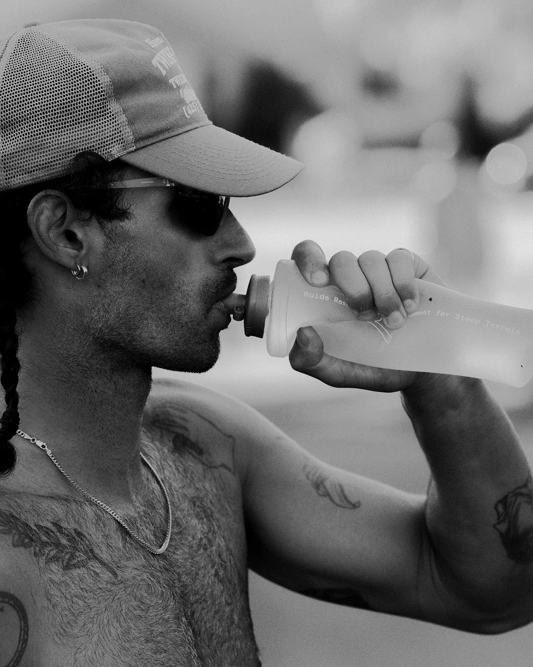 A shirtless man wearing a cap, sunglasses, and an earring drinks from a sports bottle. The image is in black and white.