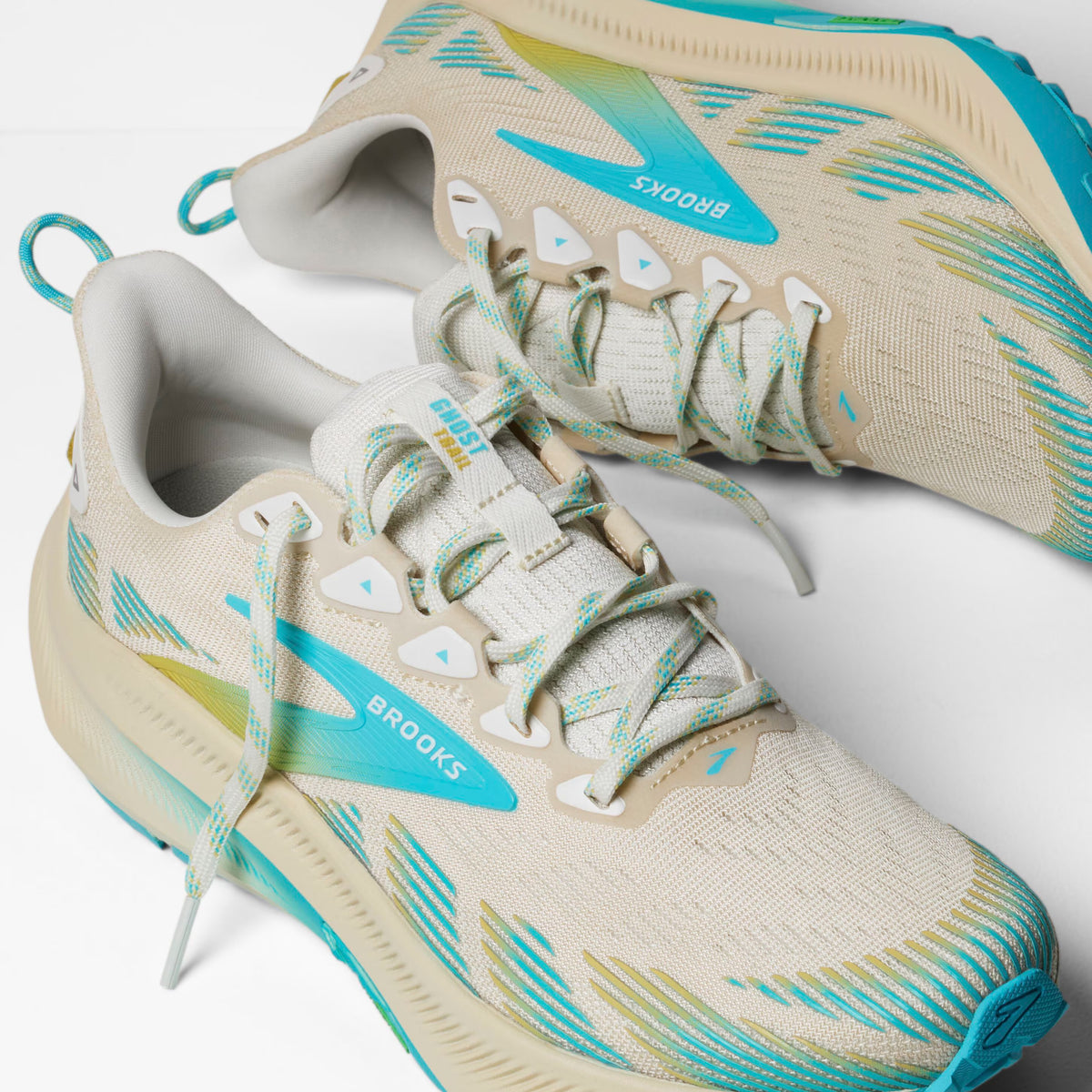 Beige and teal Brooks running shoes with white laces and blue accents are placed on a white surface, showing both the side and top views of the shoes.