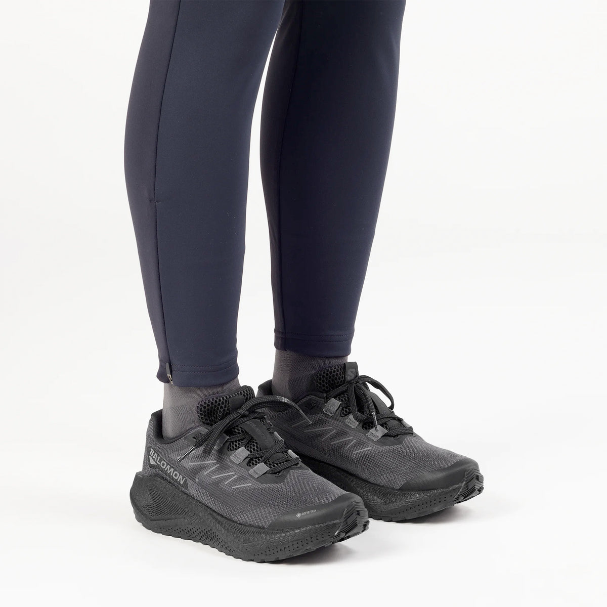 Person wearing black athletic shoes and dark leggings, standing on a white background. The shoes have thick soles and laces, and a logo is visible on the side of the shoes.