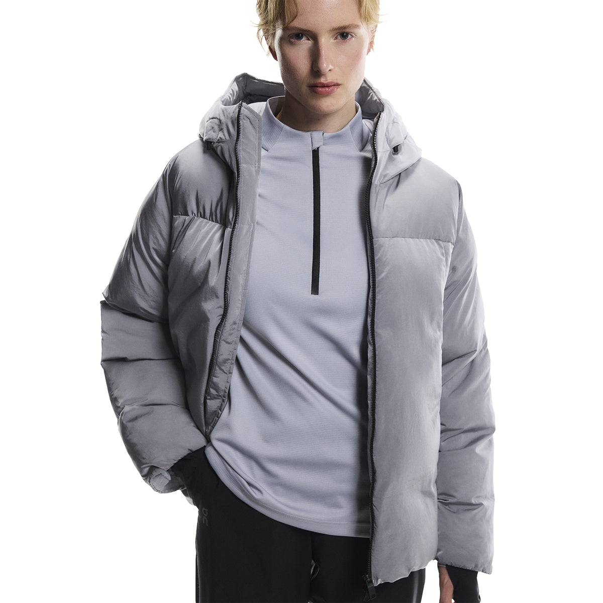 Person wearing a light gray puffer jacket over a zip-up shirt, standing against a white background with hands in pockets, looking straight ahead.