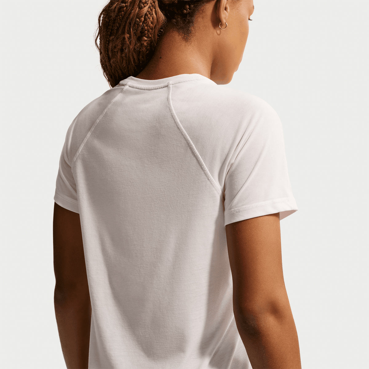 A person with braided hair is shown from the back, wearing the Nike Women’s ACG Dri-FIT Trail Running Top in Phantom / Summit White, a short-sleeved trail running shirt featuring moisture-wicking Dri-FIT technology, set against a light background.