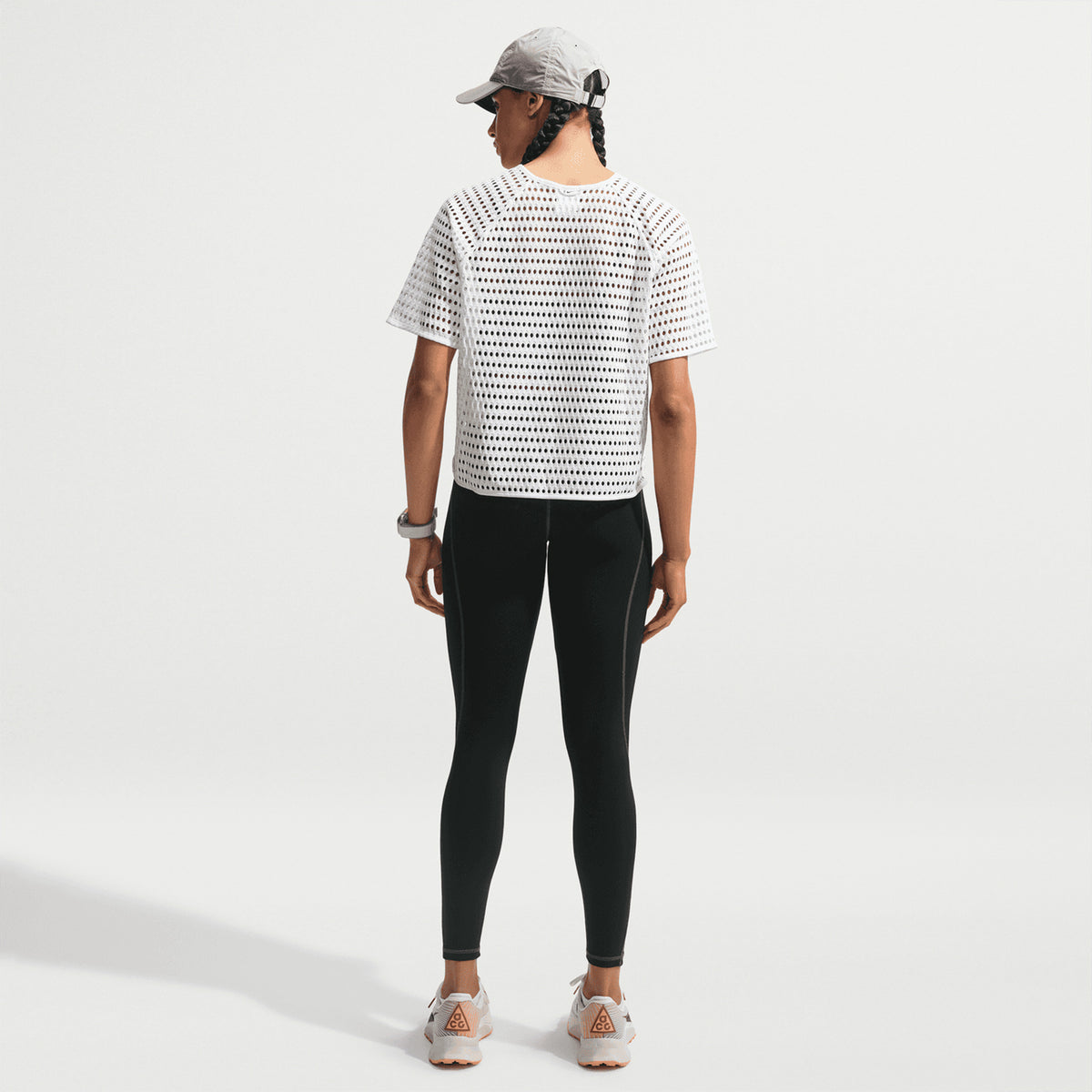 Person standing with their back to the camera, wearing a gray cap, Nike Women’s ACG Radical AirFlow Tee White / Anthracite, black leggings, white sandals, and a watch against a plain white background.