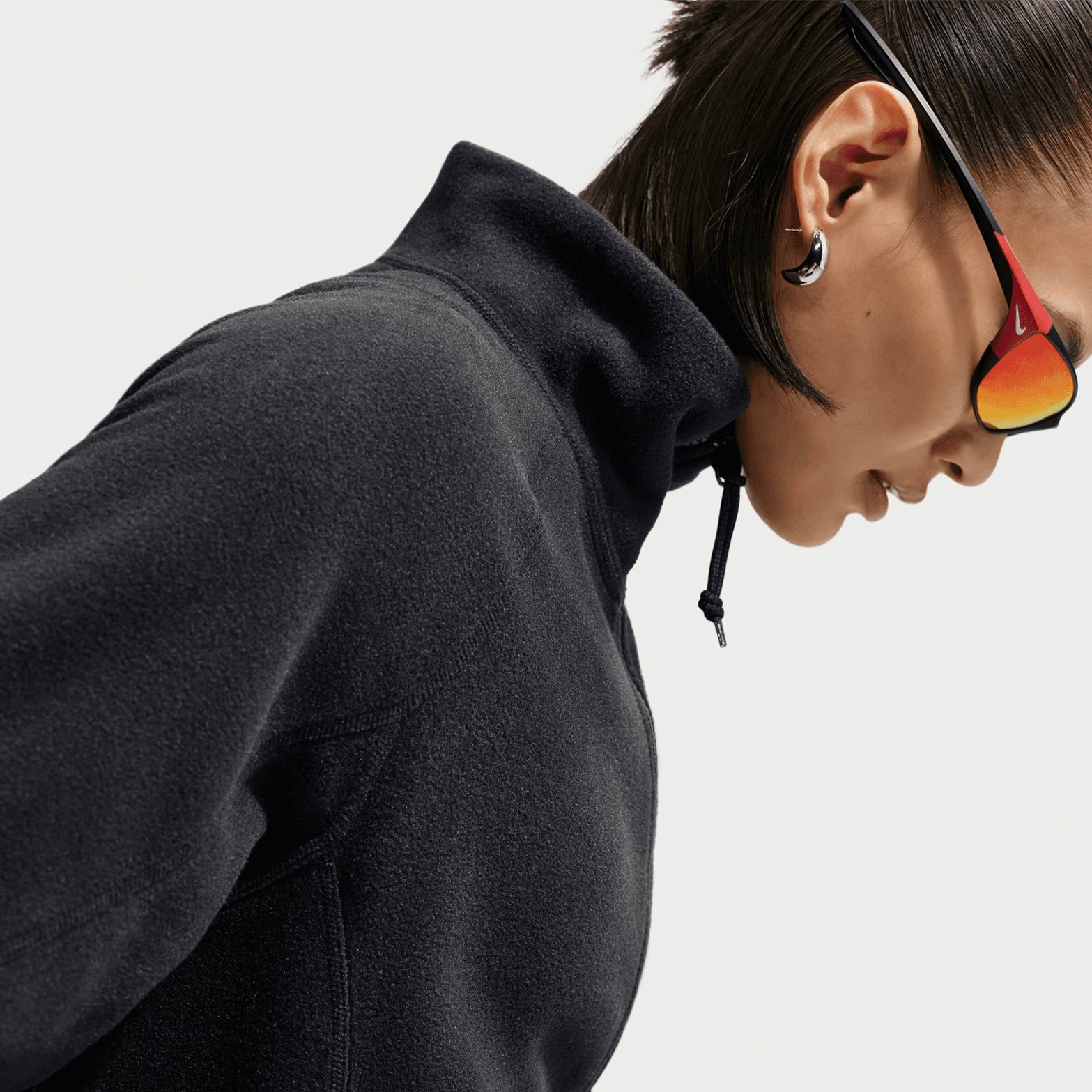 A person wearing the Nike Women’s ACG Wolf Tree 1/2 Zip Top in Black / Anthracite / Summit White and reflective sunglasses looks downward. They have short dark hair, visible hoop earrings, and a high collar with a drawstring. The background is plain white.