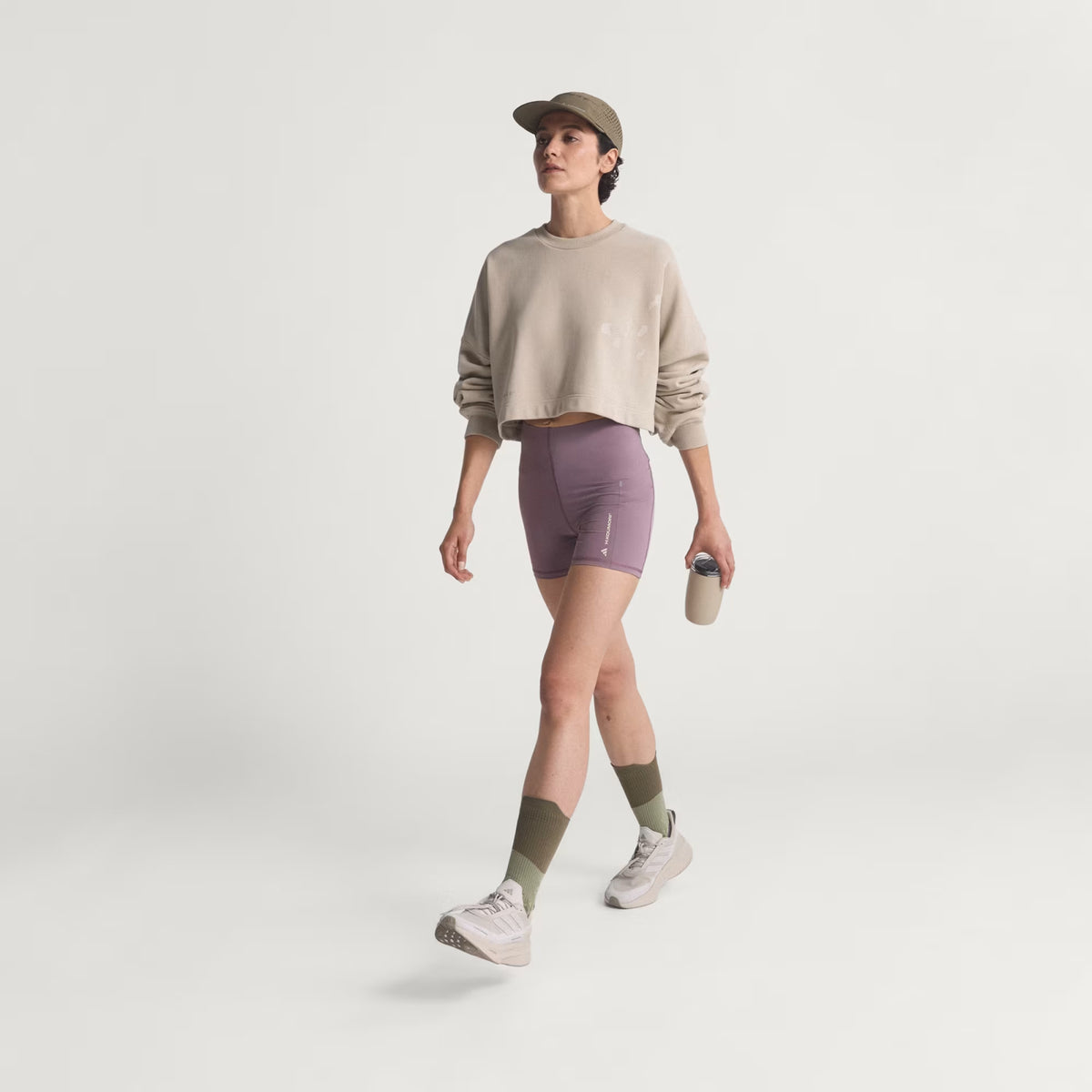 Wearing an Adidas Women’s Adi365 H.Koumori Running Crew Top in Putty Grey, a person in a beige cap, purple shorts, green socks, and white sneakers walks while holding a reusable cup. This look combines the crewneck streetwear style of the Adi365 top with performance lifestyle comfort against a plain white background.