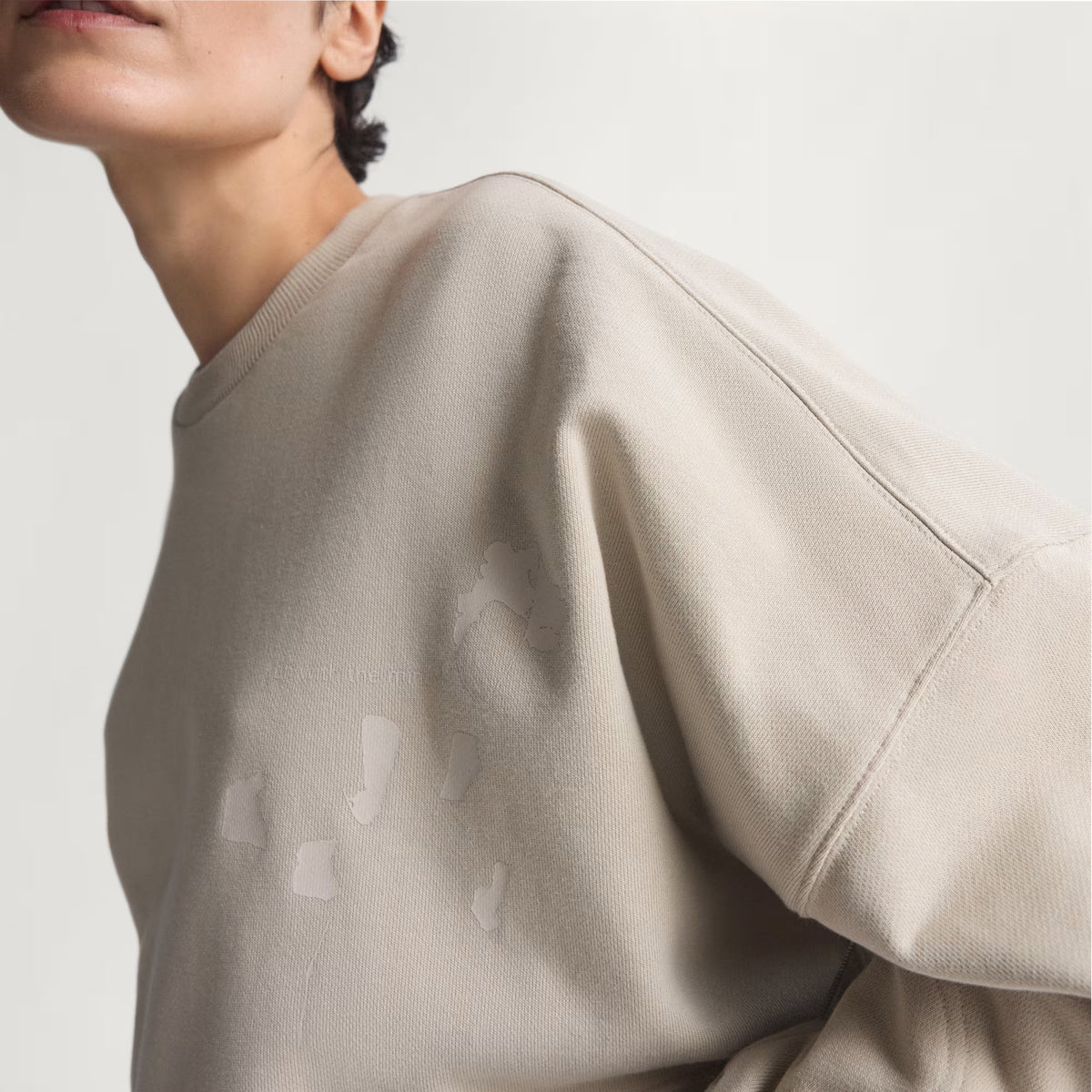 A woman wearing the Adidas Women’s Adi365 H.Koumori Running Crew Top in Putty Grey is shown from the shoulders up against a plain light background, leaning slightly forward with her face partially out of frame.