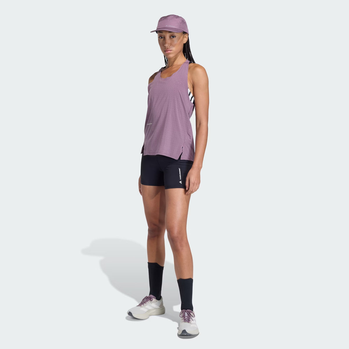 A woman wearing a purple cap, sleeveless purple top, Adidas Women’s Adi365 H.Koumori Running Short Tights in black with a high-rise waistband, black knee-high socks, and white sneakers stands facing forward against a light background.
