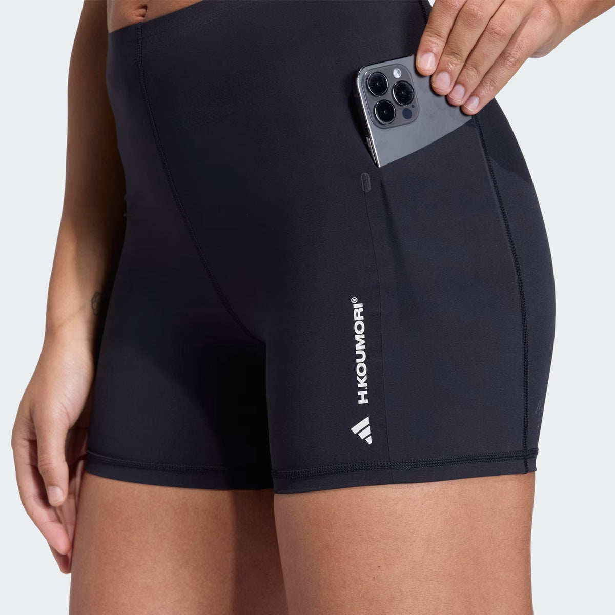 A person wears the Women’s Adi365 H.Koumori Running Short Tights in black by Adidas, featuring a high-rise waistband and convenient side pocket for a phone. The shorts display the H.KOUMORI logo in white on the left thigh, combining comfort and style similar to running leggings.
