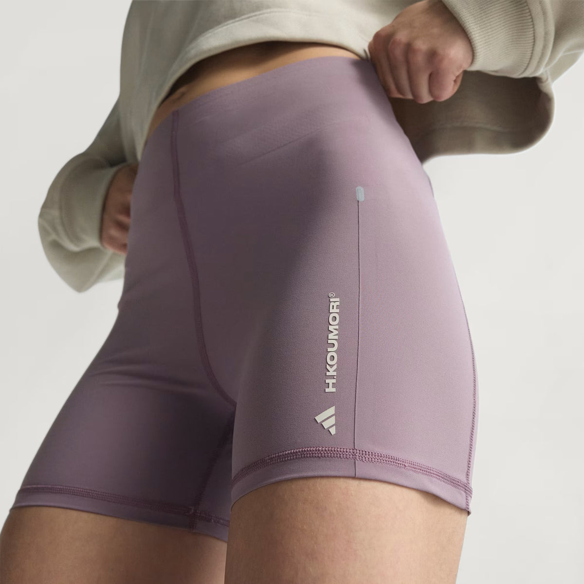 A person is wearing the Adidas Women’s Adi365 H.Koumori Running Short Tights in Shadow Fig—high-waisted, light purple athletic shorts featuring a high-rise waistband and the Hikoumori logo on the left thigh—paired with a beige cropped sweatshirt. Reminiscent of Adidas Short Leggings, these shorts also include an antimicrobial gusset for added comfort. The background is neutral and plain.