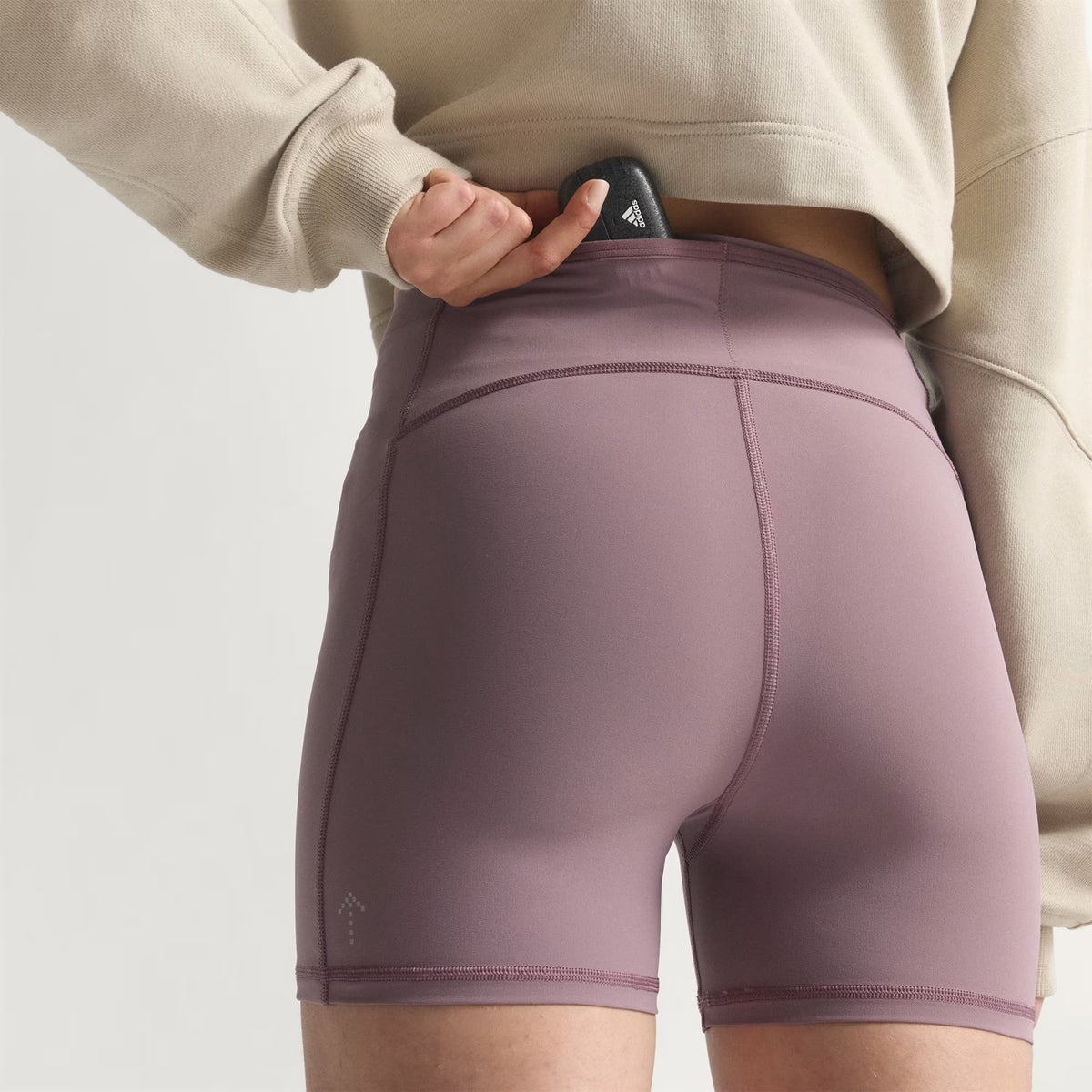A person wearing the Women’s Adi365 H.Koumori Running Short Tights in Shadow Fig by Adidas, which feature a high-rise waistband, places a black phone with a logo into the back pocket. They are also dressed in a light beige sweatshirt, set against a plain, light-colored background.