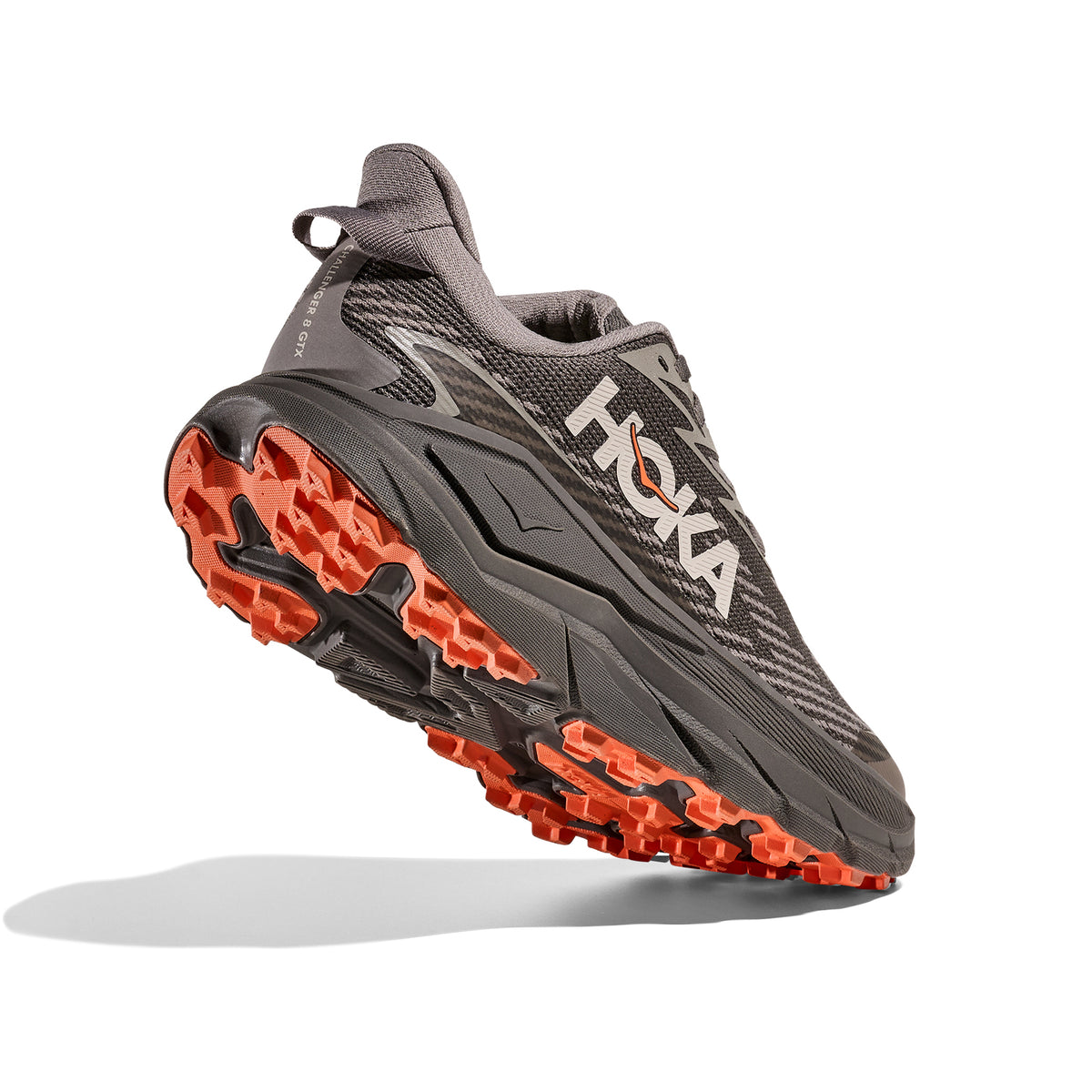 A gray Hoka running shoe with orange treads is shown at an angle, highlighting its sole and design for trail running.