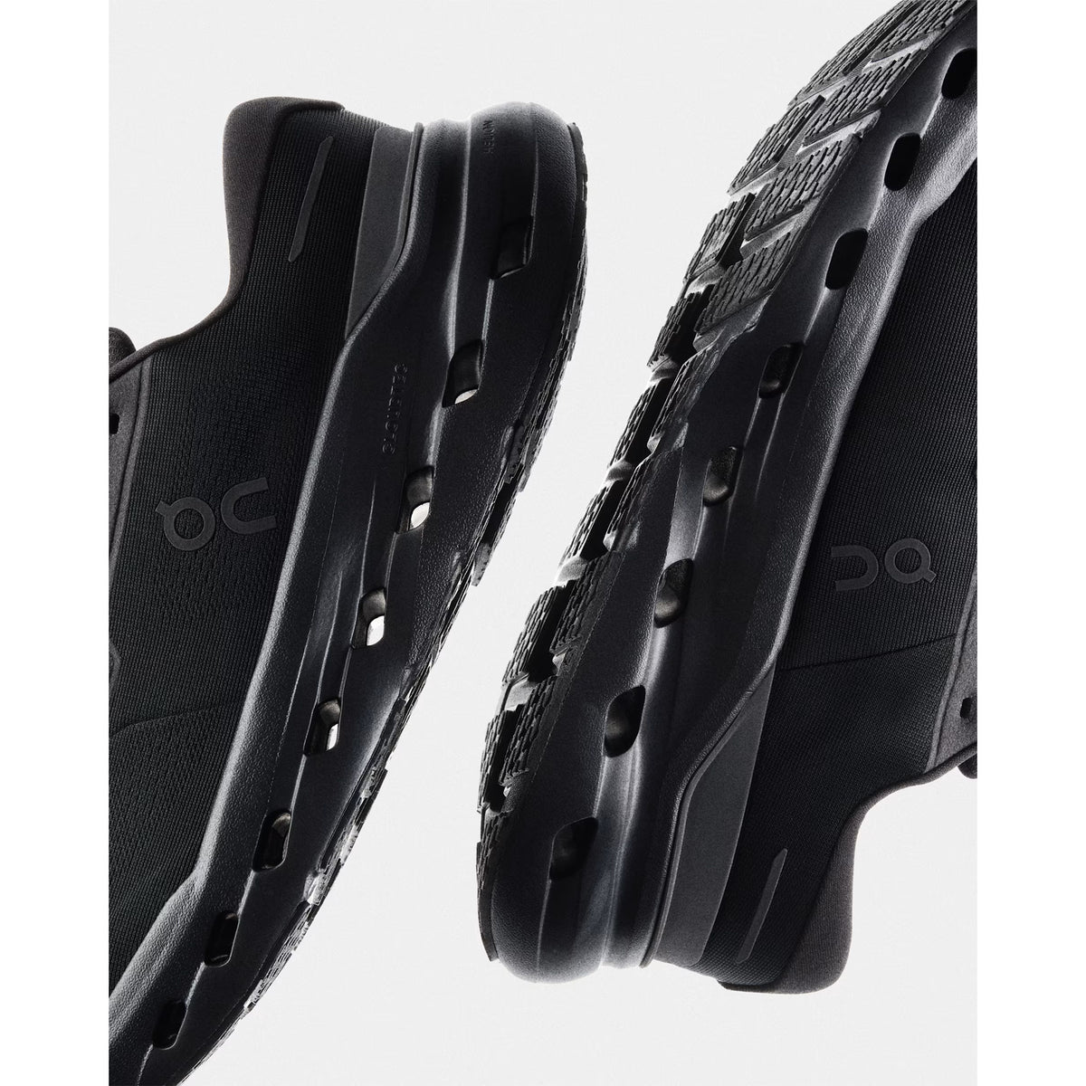 Close-up of two black sneakers facing each other, showing side and bottom views with visible logo and distinctive cut-out details along the midsole and outsole.