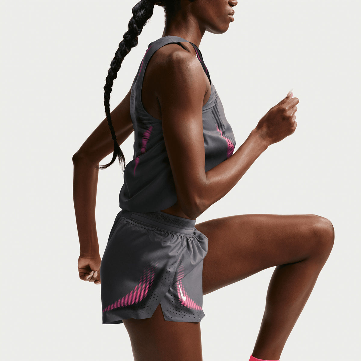 A woman wearing a gray athletic tank top and shorts with pink accents lifts her knee as if running or exercising, shown from the side against a plain white background.