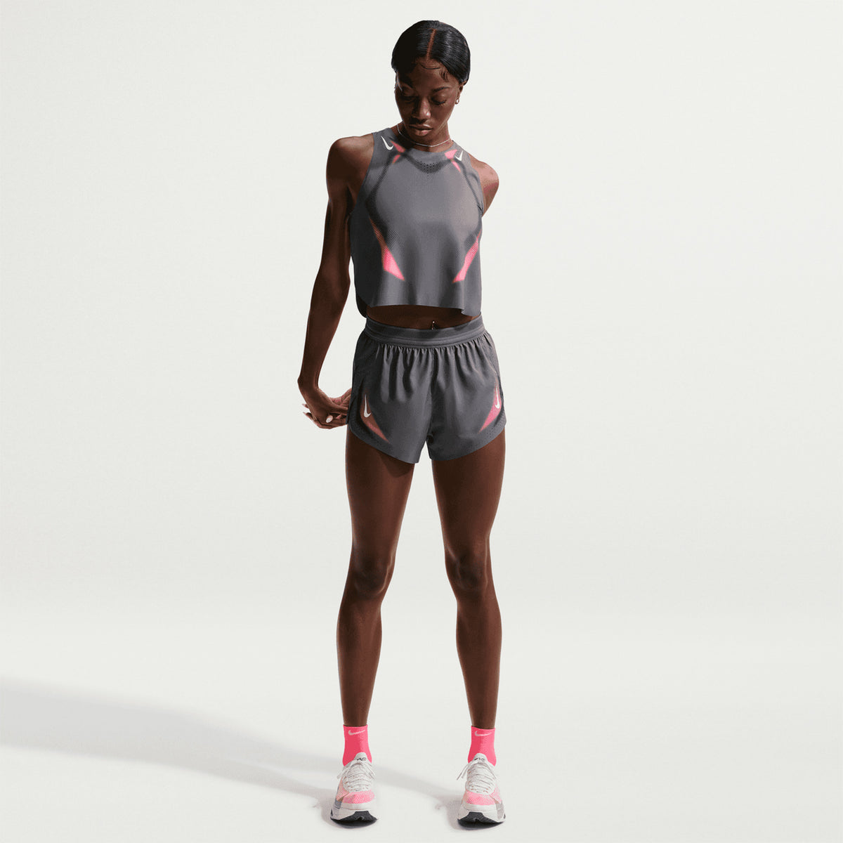 A woman wearing Nike’s Women’s Dri-FIT Aeroswift ADV 3 Inch Shorts in Iron Grey and White stands against a plain white background, looking down as she holds her lightweight running shorts. Pink-accented athletic wear, pink socks, and white sneakers complete her outfit.