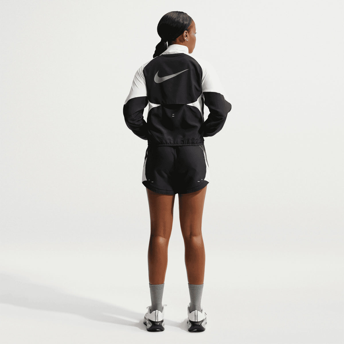 A person stands facing away, wearing the Nike Women’s Dri-FIT Tempo Swoosh 1/2 Zip Running Jacket in Black, White, and Cool Grey with shorts, gray socks, and white sneakers, on a plain white background.