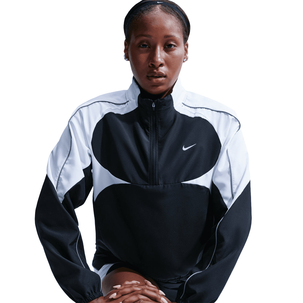 A woman wearing the Nike Women’s Dri-FIT Tempo Swoosh 1/2 Zip Running Jacket in Black, White, and Cool Grey sits with her hands resting in front of her, looking directly at the camera against a plain white background.