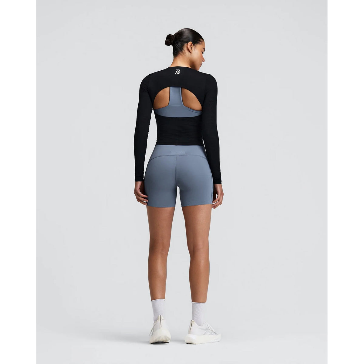 A woman stands facing away, wearing the Bandit Women’s GeoMesh Eclipse Long Sleeve Top in black, featuring a back cutout, paired with grey athletic shorts, white socks, and white sneakers against a plain light background.