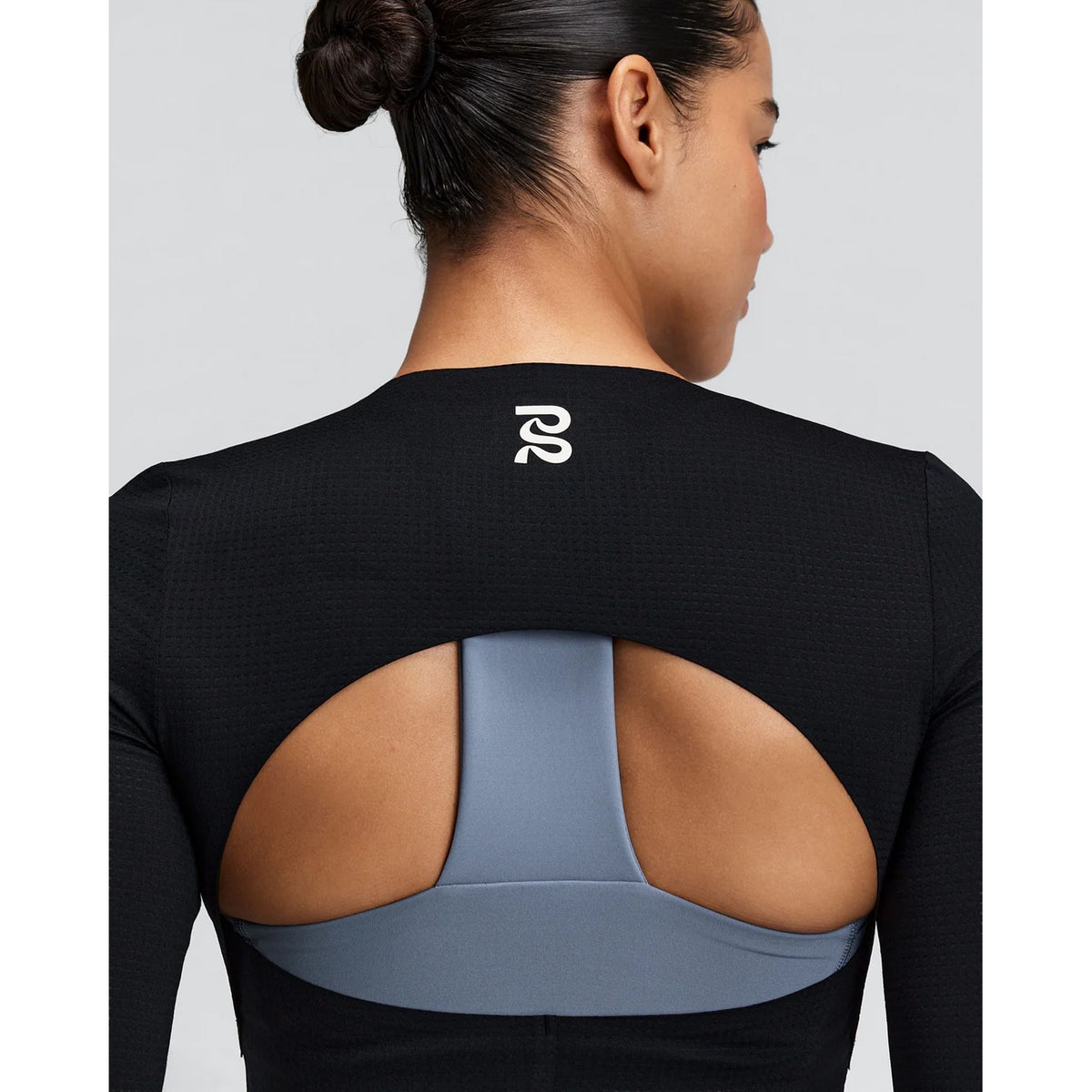 A woman with dark hair in a bun wears the Bandit Women’s GeoMesh Eclipse Long Sleeve Top in black, which features an open-back design and breathable mesh long sleeves, revealing a light blue sports bra underneath. The top displays a white logo on the upper back.
