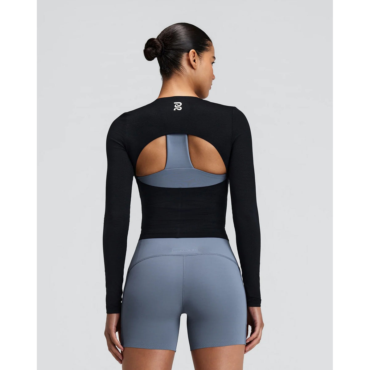 A woman stands facing away, wearing the Bandit Women’s GeoMesh Eclipse Long Sleeve Top in black, which features an open back cutout and is designed with advanced moisture management and breathable mesh construction. She pairs it with high-waisted gray bike shorts, while her hair is tied back neatly in a bun.
