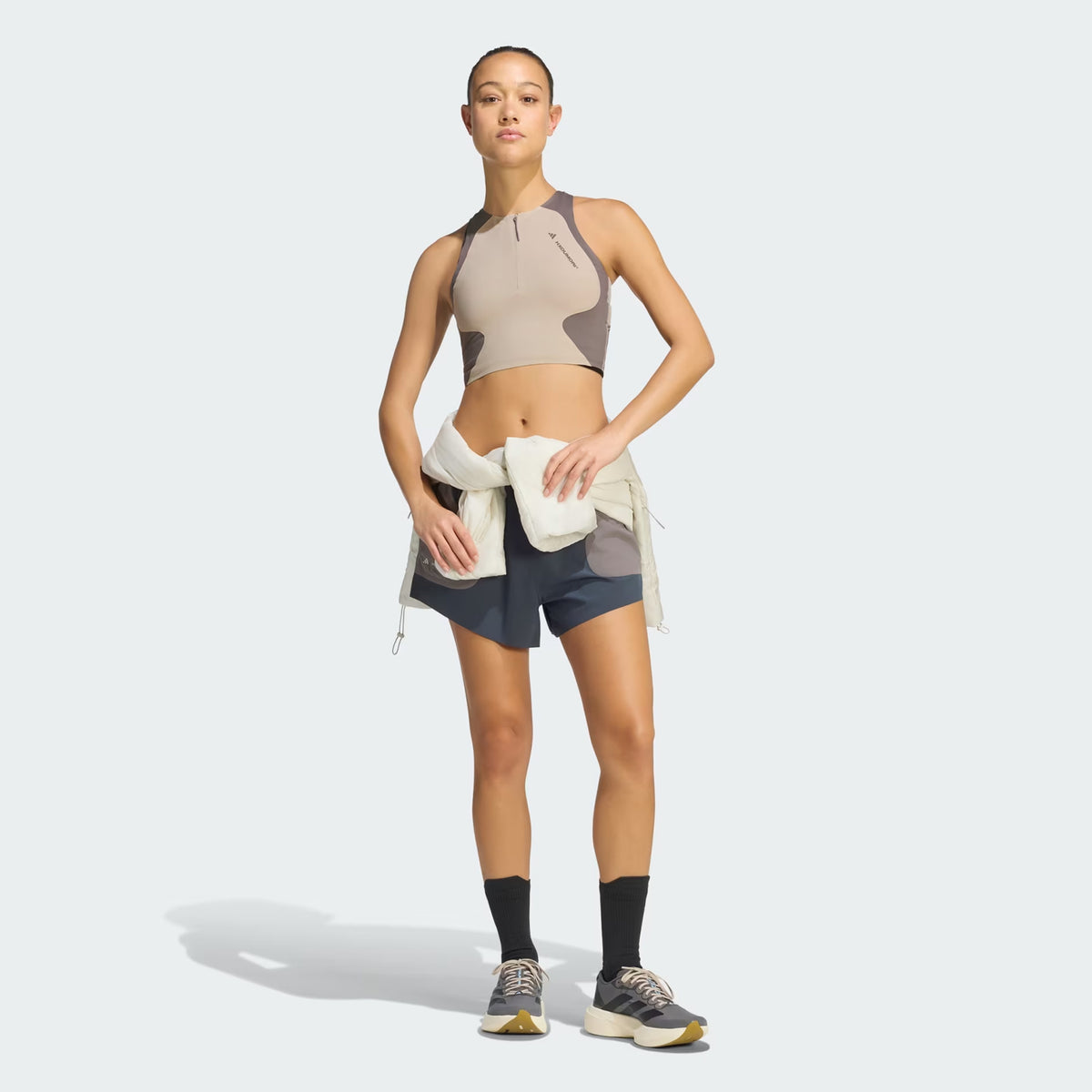 A woman stands against a plain light background wearing the Adidas Women’s Hermanos Koumori 1/2 Zip Running Crop in Wonder Beige / Charcoal, paired with blue shorts, black socks, sneakers, and a light jacket tied around her waist.