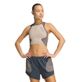 A woman with braided hair stands against a white background, hands on hips and looking to the side, wearing the Adidas Women’s Hermanos Koumori 1/2 Zip Running Crop in Wonder Beige and Charcoal paired with dark athletic shorts.