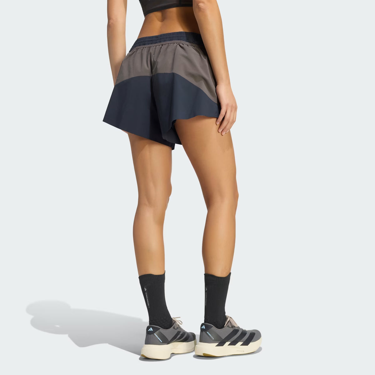 A woman stands facing away, wearing a black sports bra, Adidas Women’s Hermanos Koumori 3 Inch Running Shorts in Night Grey / Charcoal, black socks, and gray sneakers against a plain light background.