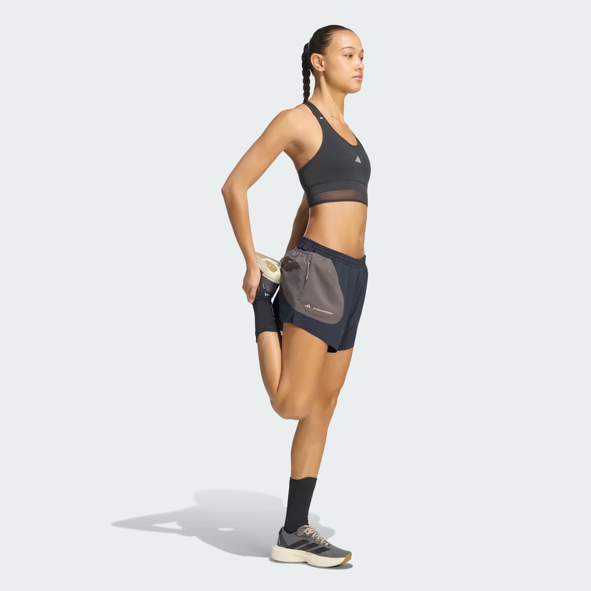 A woman in athletic wear stands on one leg, stretching her other leg by holding her foot behind her. She is wearing a sports bra, performance running gear including Adidas Women’s Hermanos Koumori 3 Inch Running Shorts in Night Grey / Charcoal, along with socks and sneakers, against a light background.