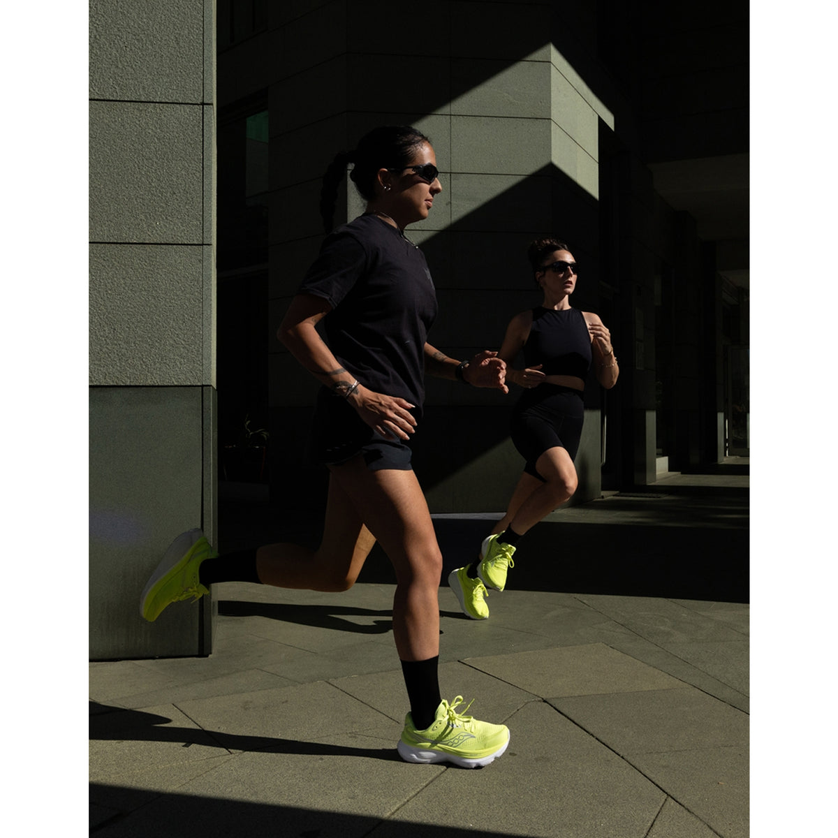 Two people wearing black outfits and bright yellow sneakers run side by side on a city sidewalk, shaded by tall buildings.