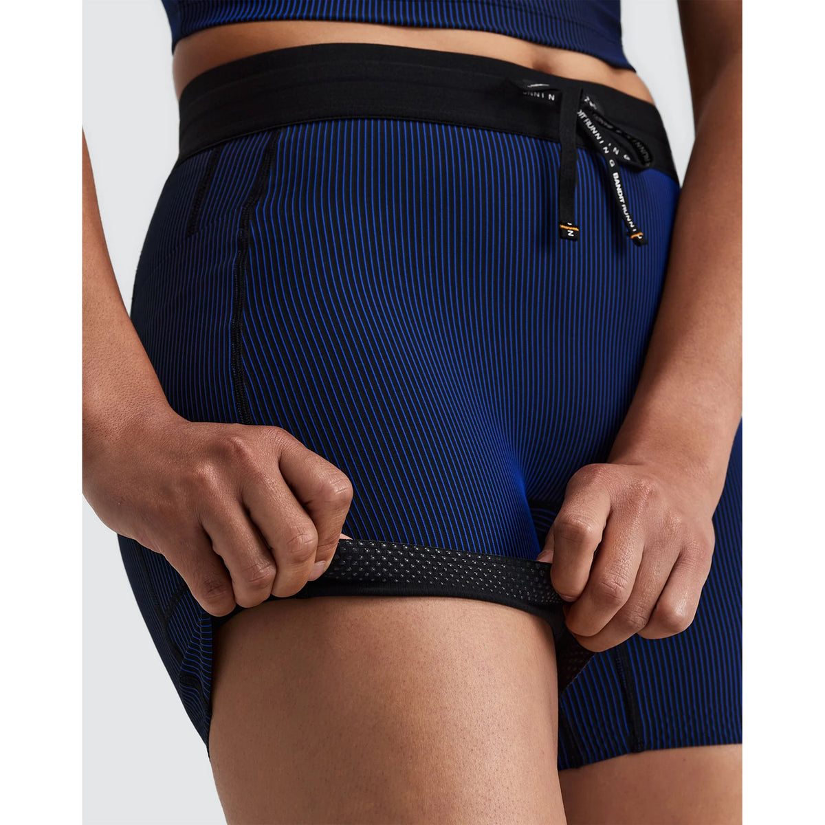 Close-up of Women’s Striped Cadence 5″ Compression Shorts Black / Bauhaus Blue held in a person’s hands, showing the black and blue striped ultra-compressive fabric, black waistband, and perforated inner leg gripper.