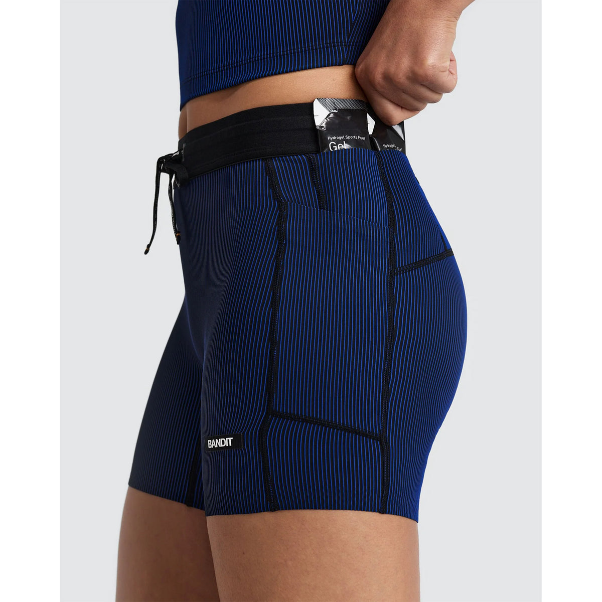 A person wearing Bandit Women’s Striped Cadence 5″ Compression Shorts Black / Bauhaus Blue, featuring blue compression material with black vertical stripes, a drawstring waistband, and 5-pocket storage, including a side pocket holding a gel packet.