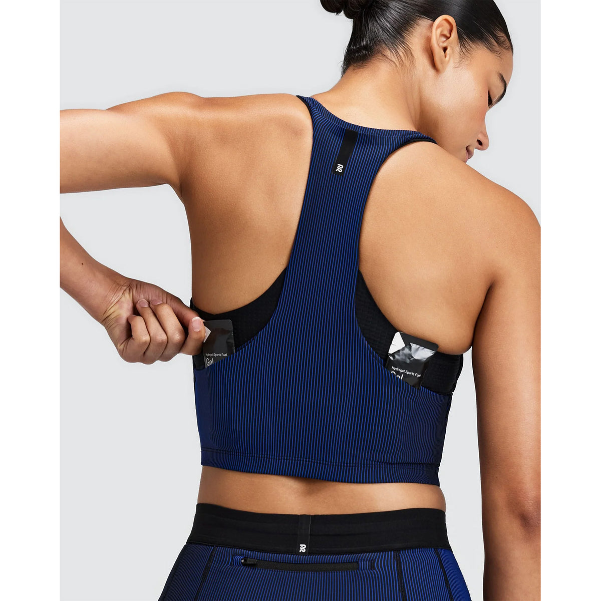 A person wearing a Women’s Striped Cadence Zip Front T-Back Race Crop Black / Bauhaus Blue shown from the back, tucking a sports gel packet into one of the pockets of its seven-pocket system.