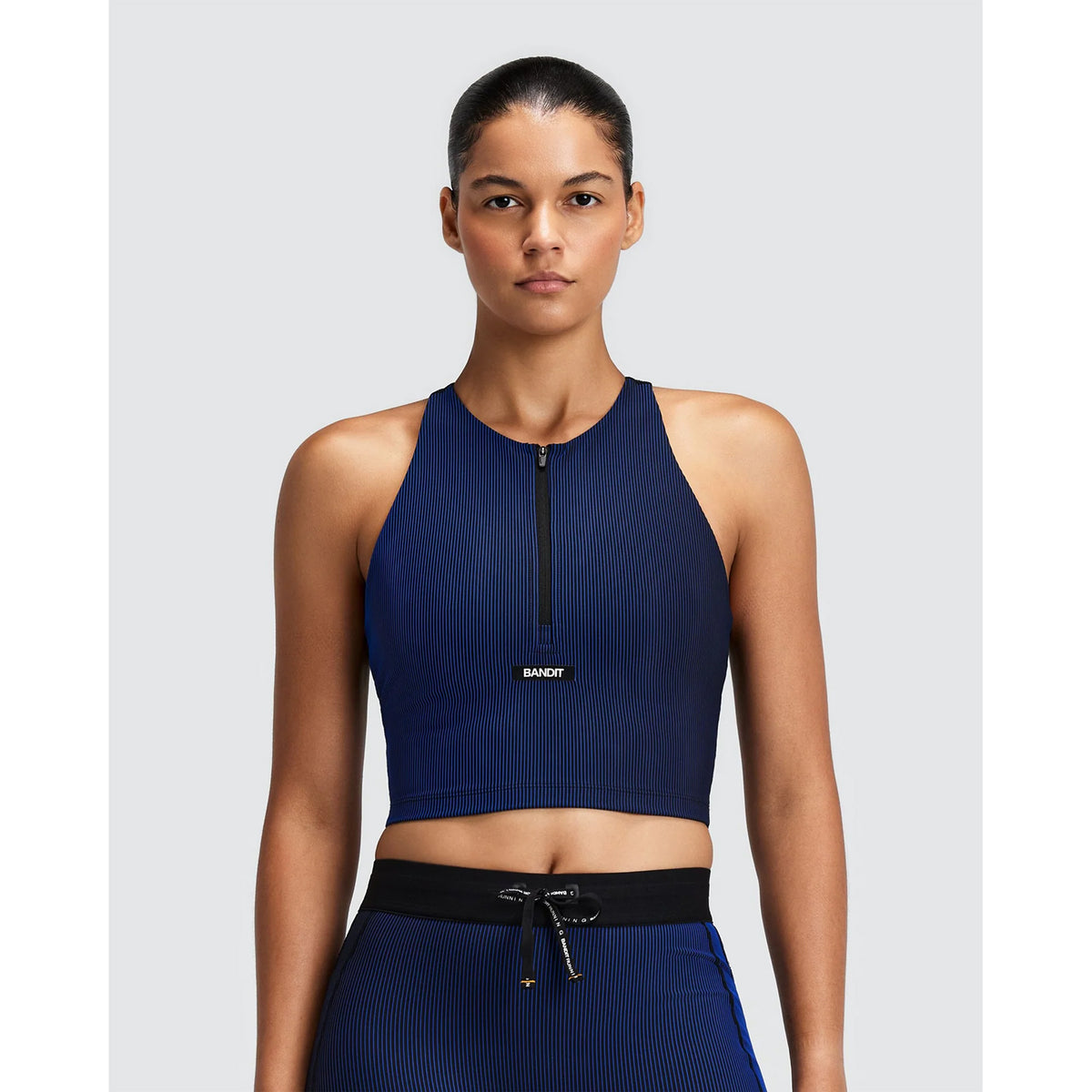 A woman wears the Bandit Women’s Striped Cadence Zip Front T-Back Race Crop in Bauhaus Blue, a dark blue, ribbed compression, zip-front cropped tank top with a T-back design.
