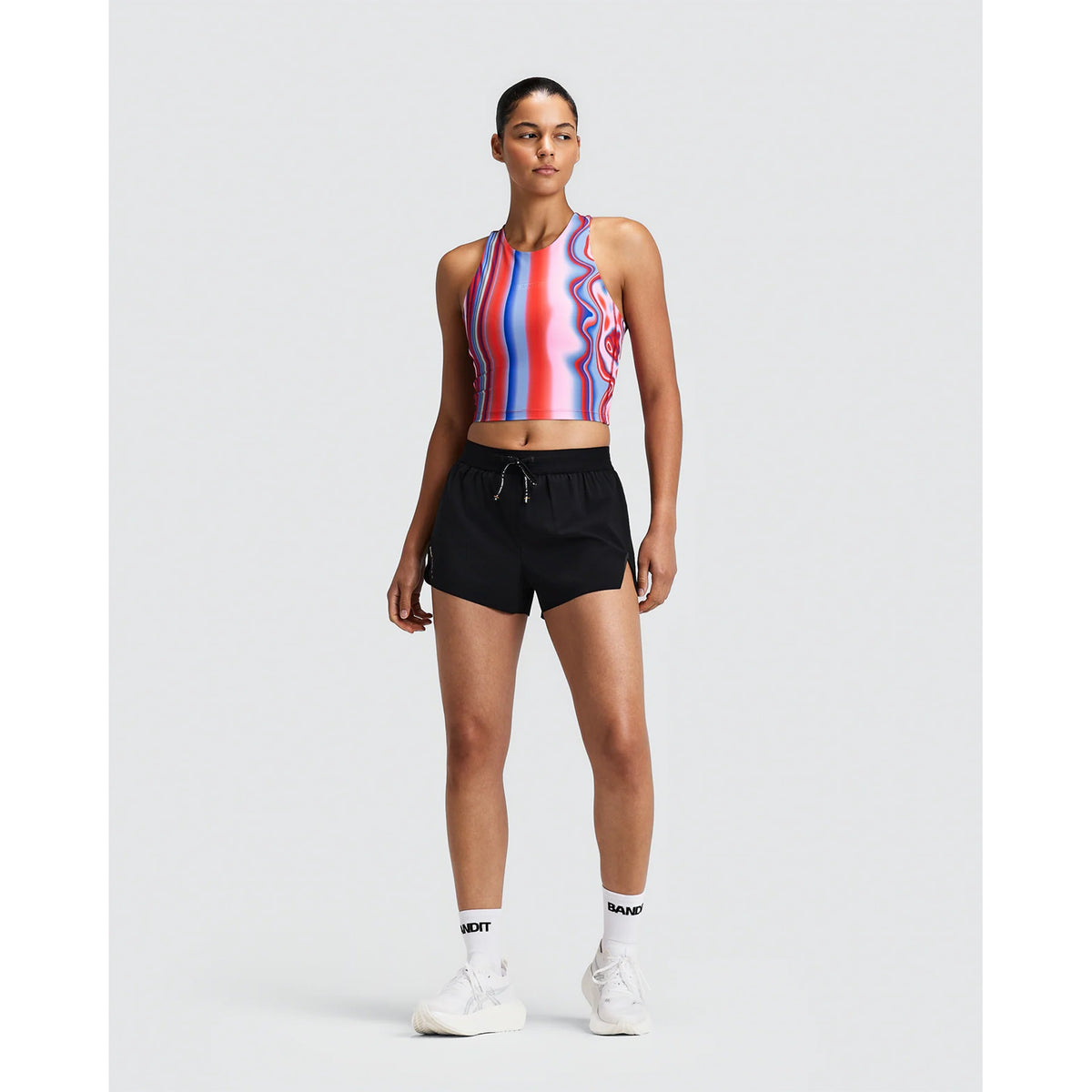 A person wears a Bandit Women’s Vanisher Race Crop Melted Stripe Gradient top with a red, blue, and white vertical wavy stripe pattern, paired with black athletic shorts featuring a seven-pocket system, white crew socks, and white sneakers.