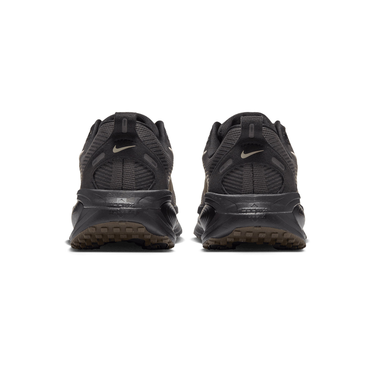 The back view of a pair of Women’s Vomero 18 Running Shoes in Medium Ash / Dark Hazel / Black / Desert Khaki, featuring dark-colored uppers, textured midsoles, and brown lugged outsoles.