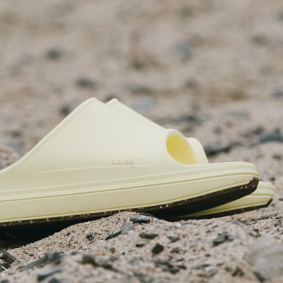 A pair of light yellow slide sandals resting on a sandy, rocky surface.