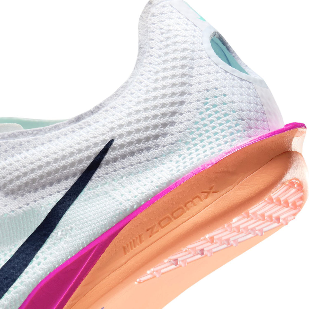 Close-up of a white athletic shoe with a mesh upper, black swoosh, and a thick sole featuring pink, peach, and orange accents. The sole has spikes and NIKE ZOOMX embossed on the side.