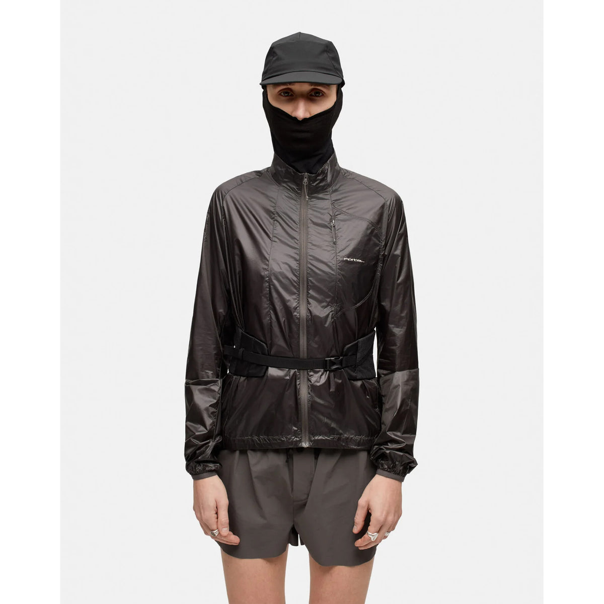 A person wearing a black cap, face covering, and an abrasion-resistant shiny black jacket with a belt, paired with gray shorts and the Portal Borders Cargo Belt Black Beauty hip pack for secure storage, stands against a plain white background.