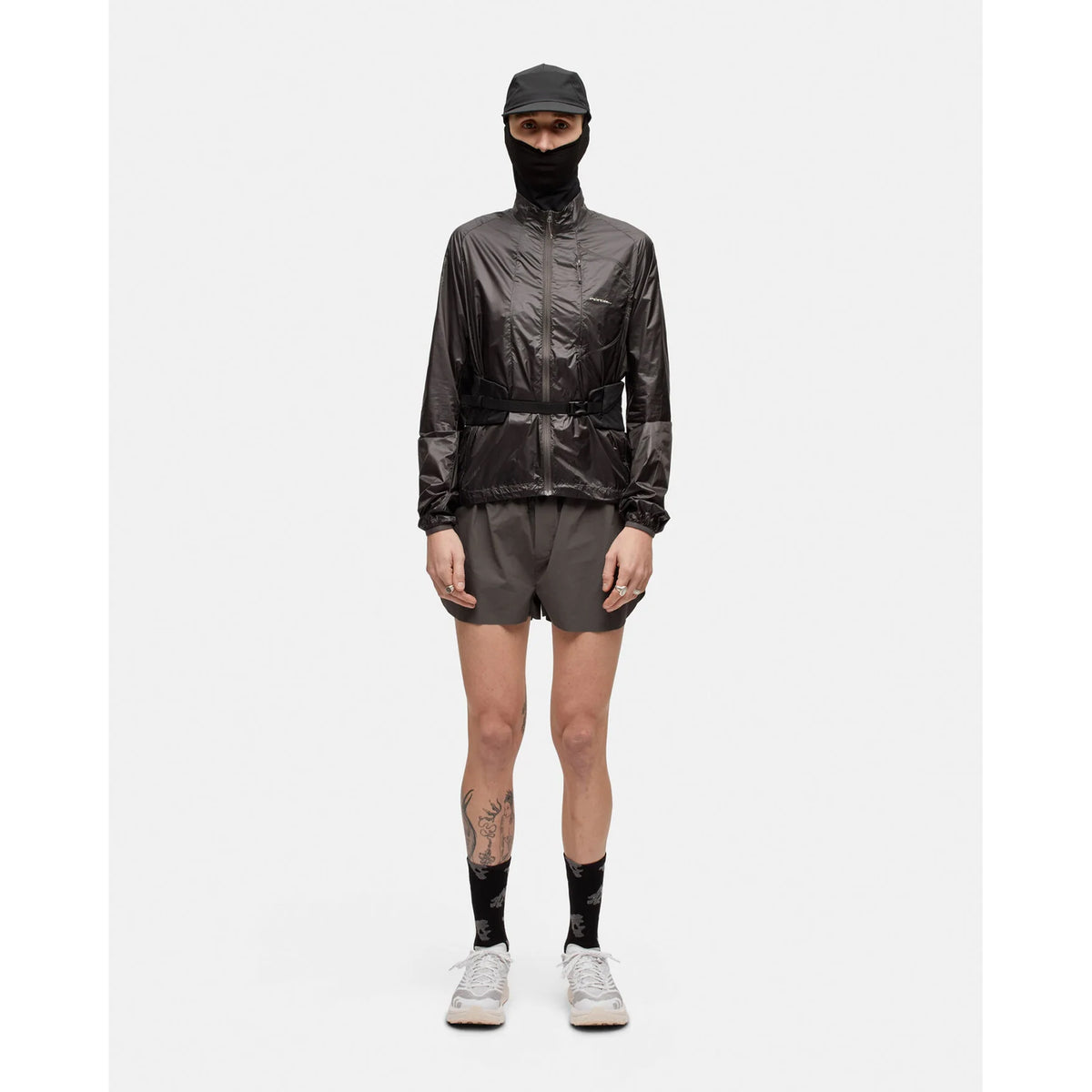 Standing against a white background, the person is wearing a black cap, black jacket, black face covering, gray shorts, and the Portal Borders Cargo Belt in Black Beauty for secure storage. The outfit is finished with black socks featuring white designs and abrasion-resistant white running shoes. A tattoo is visible on one leg.