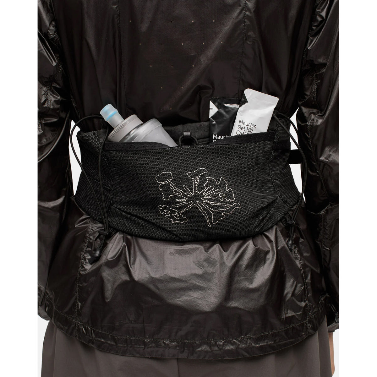A person wearing a black jacket has a Portal Borders Cargo Belt Black Beauty—a black, abrasion-resistant hip pack with a floral design that provides secure storage for a water bottle, an energy gel, and snack packets.
