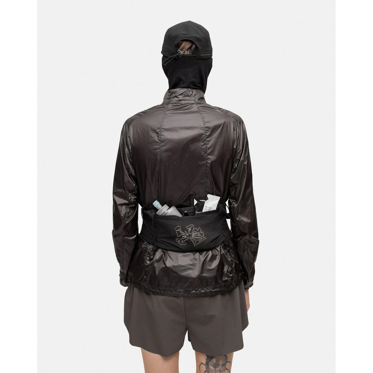 Person in a shiny black, abrasion-resistant jacket, shorts, and cap, facing away. They wear the Portal Borders Cargo Belt Black Beauty hip pack, which provides secure storage for various items. Tattoo visible on back of left thigh. Background is plain white.
