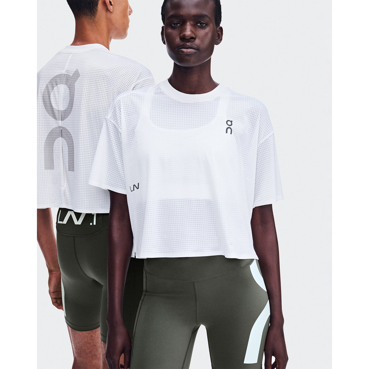 Two people stand together against a white background, both dressed in athletic wear from Lane 1 Collective. The person in front wears the Cropped Run-T LN1 White by On—a lightweight, white crop top with back ventilation—paired with olive green leggings. Behind, another person wears a white top and dark leggings. Both tops feature subtle branding.