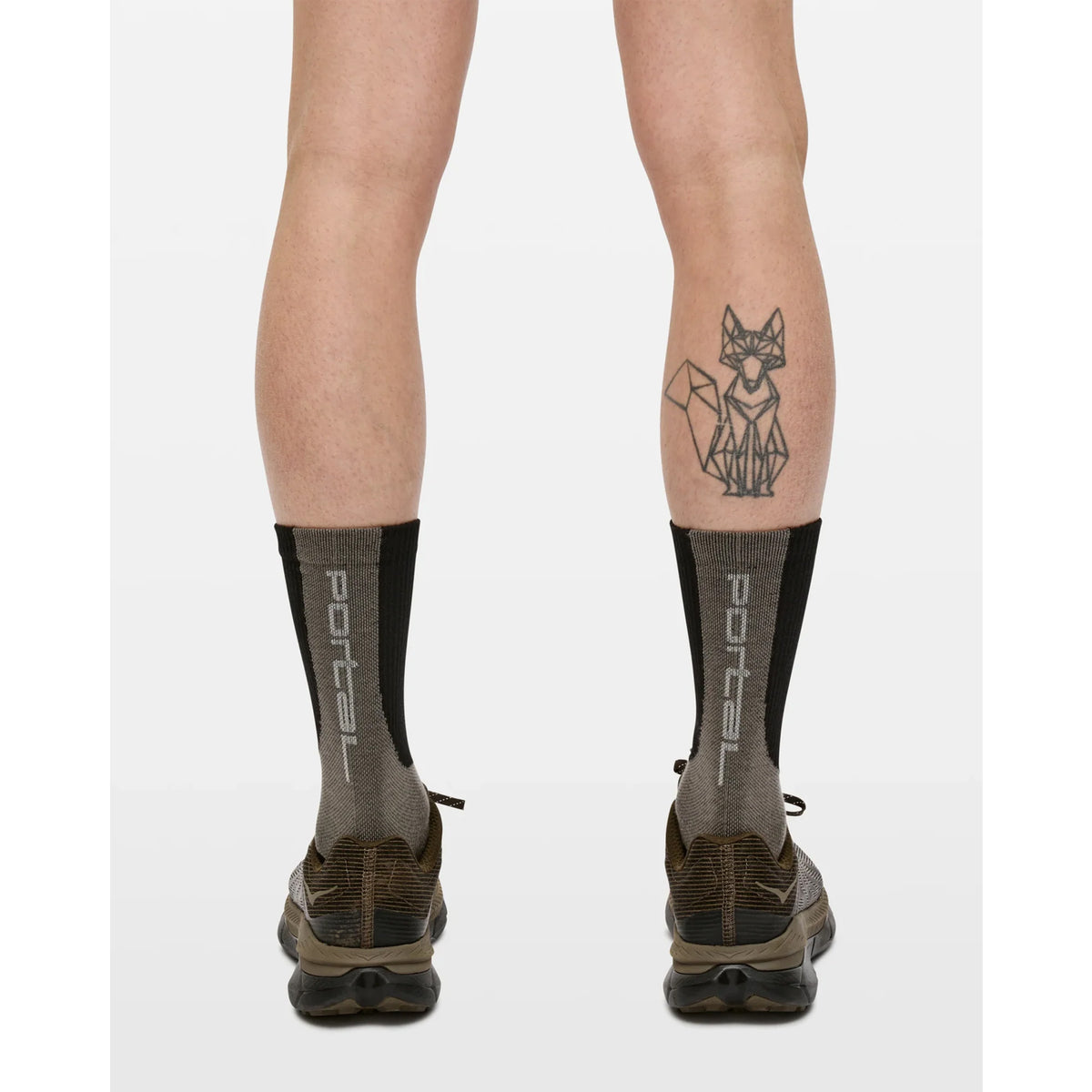 A person stands with their back to the camera, wearing Portal Lineage All Purpose Socks in Black Beauty—cotton blend socks designed for all-purpose active use—with brown shoes. A geometric fox tattoo is visible on the left calf. The background is plain white.